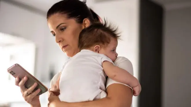 žena sa bebom u naručju drži u ruci mobilni telefon