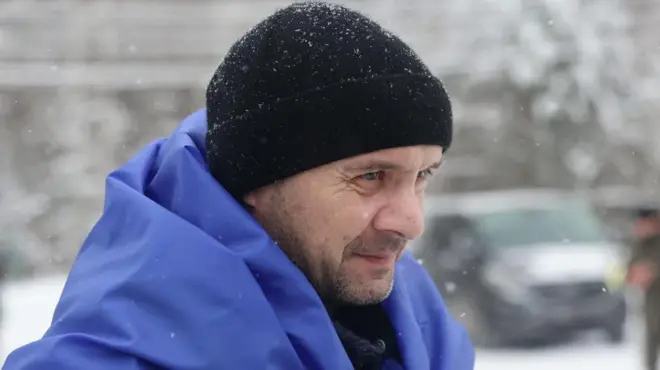 Чоловік у чорній шапці з синім прапором на плечах.