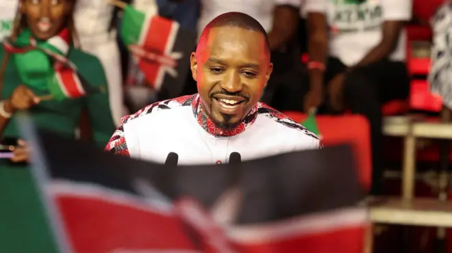 Boniface Mwangi yambaye ishati y'umweru n'amabara y'umukara n'umutuku, afashe ibendera rya Kenya