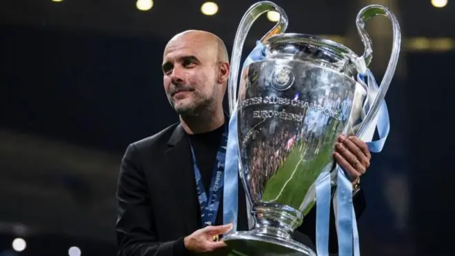 المدرب الإسباني بيب غوارديولا يحمل كأس دوري أبطال أوروبا - فاز به فريقه مانشستر سيتي 2023
يرتدي سترة سوداء ويحمل كأسا فضية تتدلى منها شرائط زرقاء - ويضع ميدالية ذهبية