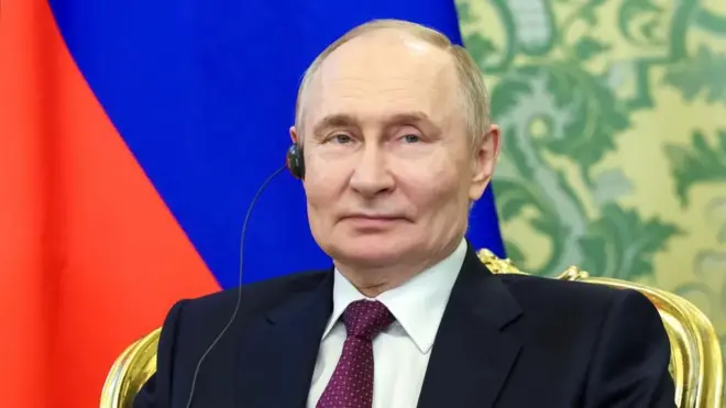 А хеадсхот портраит оф Владимир Путин смилинг слигхтлy агаинст тхе ред анд блуе бацкдроп оф а флаг