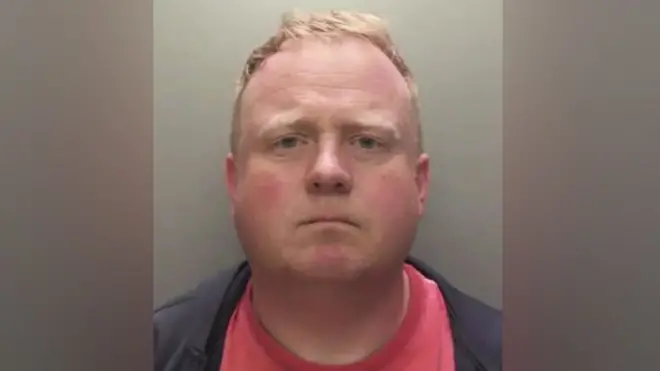 Robert Bush wit short, light brown hair show for dis straight‑on mugshot foto. Im no dey smile or frown, im wear dark jacket ontop red shirt. Di background dey plain and grey.
