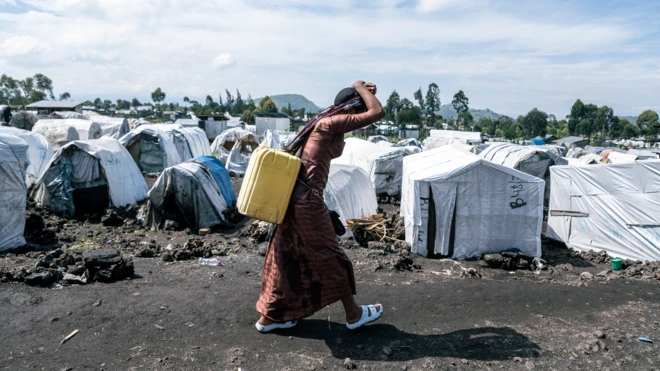 Umugore wahunze uhetse ijerikani y'amazi ayishumikishije igitenge kizingiye ku mutwe, agiye aho aba mu nkambi ya Shabindu, mu nkengero ya Goma, muri DR Congo, ku itariki ya 9 Mata (4) mu mwaka wa 2024
