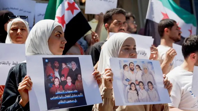 Önde iki kadın ailesinin fotoğraflarını turuyor, gerilerinde bir kadın ve birkaç adam daha var, Suriye bayrakları ve pankartlarla protesto ediyorlar.