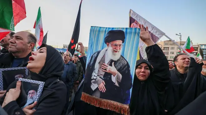 Kalabalık bir grup miting alanında. Sağ tarafta bir kadının elinde Ali Hamaney'in bir fotoğrafı var. Sol tarafta Hamaney'in fotoğrafına sarılmış ağlayan başka bir kadın var. Arkada erkekler var, bazıları ellerinde bayrak tutuyor. 