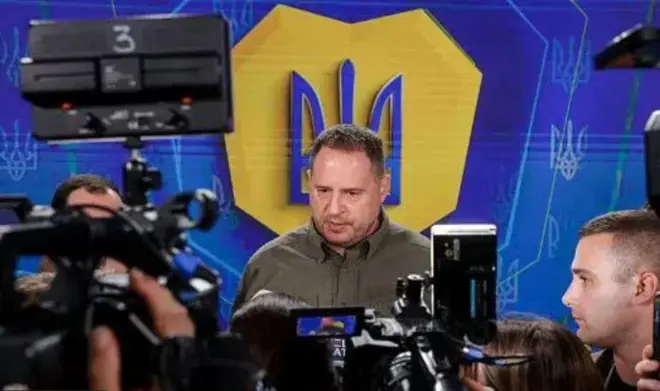 Андрій Єрмак в оливковій сорочці стоїть на синьому фоні в оточені журналістів з камерами