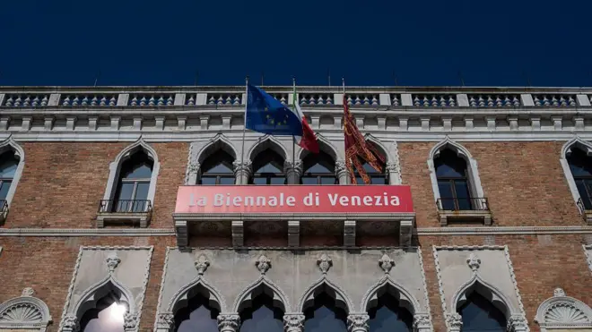 Венецианский готический фасад, украшенный баннерами La Biennale di Venezia 2026, указывающими на различные дисциплины и периоды культурной программы, с европейским, итальянским и венецианским флагами над ним, в историческом архитектурном антураже городского центра.