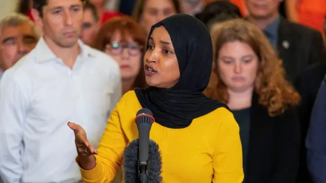 La congresista Ilhan Omar hablando ante un micrófono, con un jersey amarillo y un hiyab negro.
