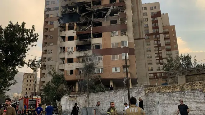 A burned hole in an apartment building as emergency workers stand on the ground below