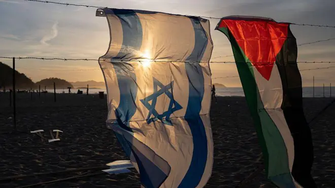 Uma bandeira israelense e outra palestina penduradas em uma instalação de arame farpado. Ao fundo, a praia de Copacabana, no Rio de Janeiro, Brasil.