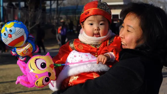中国の屋外で黒いコートを着た大人が笑いながら、伝統的な中国の衣装を着た赤ちゃんを抱いている。赤ちゃんはキャラクターものの風船を握っている