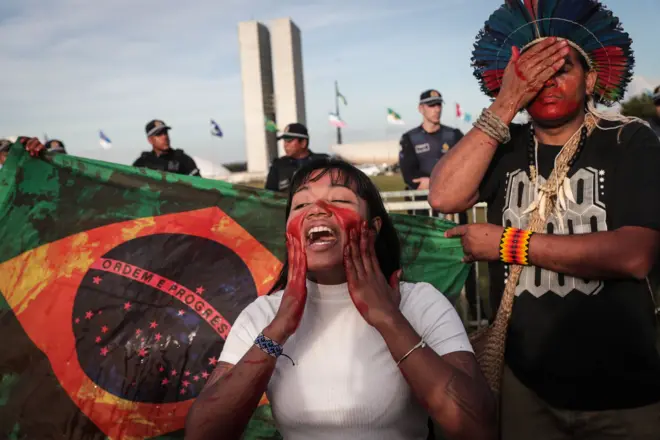 Dois indígenas protestando ao ladojoguinho que da dinheirobandeira do Brasil com aparência ensanguentada e prédios do Congresso Nacional atrás