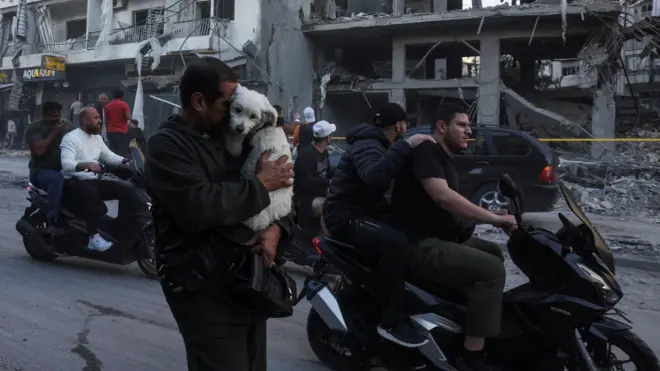 صورة لأشخاص يعبرون أمام مبنى مدمر في ضاحية بيروت الجنوبية بفعل غارات إسرائيلية. 