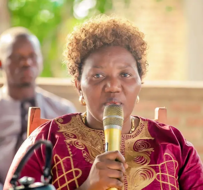 Rose Nkorerimana arimo kuvugira mu ndangururamajwi. Yambaye ikanzu iboneka igice cyo hejuru iriho ibara ritukura na khaki. Inyuma ye hari umugabo utaboneka neza.