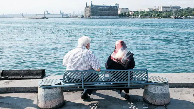 Kadıköy'de Haydarpaşa Garı'nı gören bir açıdan deniz kenarındaki banka oturmuş yaşlı bir çift. Arkadan gördüğümüz çiftten soldaki erkek olanı üzerinde kısa kollu bir gömlekle otururken sağdaki kadının üstünde ise siyah bir kıyafet ve desenli bir başörtüsü var