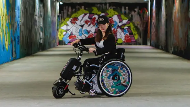 검은색 모자를 쓴 여성이 벽에 그래피티 벽화가 그려진 길 위에 휠체어를 타고 있다. 그가 탄 휠체어 바퀴에도 벽화와 비슷한 그래피티가 그려져 있다.