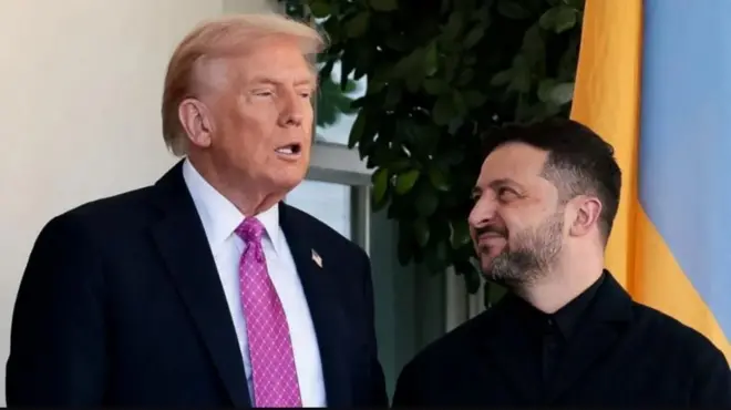 Trump mw'ikoti yirabura n'ikaruvati y'iroza ahagararanye na Zelensky yambaye ivyirabura, inyuma hari igiti n'ibendera