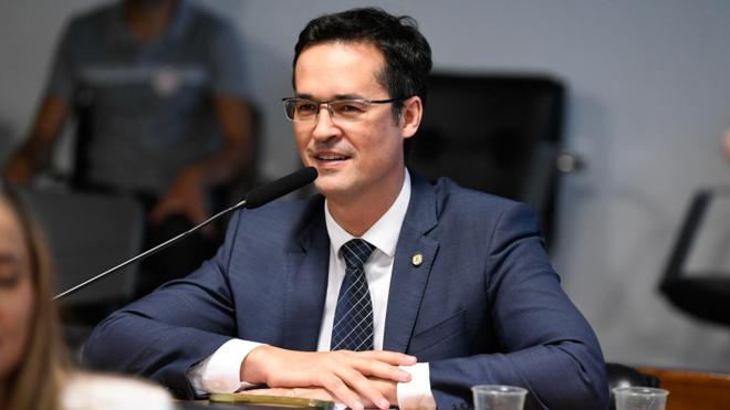 Deltan sorrindo contidamente enquanto fala, sentado diantejackpot comunitário bet365mesa no Senado 