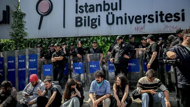 Bilgi Üniversitesi'nin girişinde oturan bir grup öğrencinin arkasında polisler kalkanlarıyla ayakta duruyor