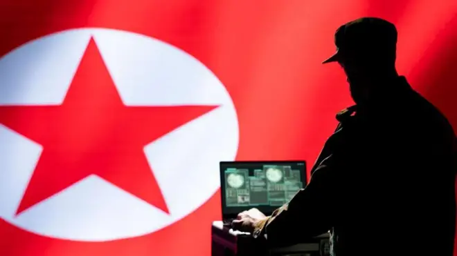 شخص يعمل على جهاز لابتوب وفي الخلفية علم كوريا الشمالية