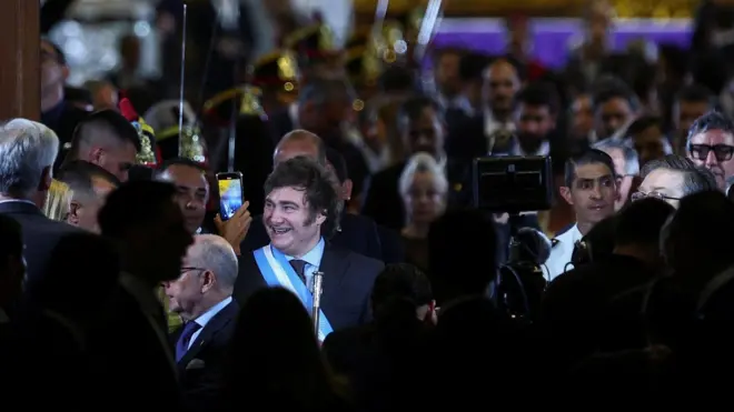 Milei sorrindo com faixa presidencial, rodeado por várias pessoas durante a posse