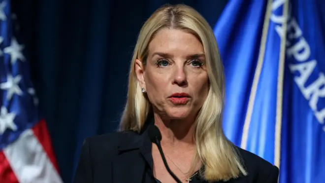 Pam Bondi has a stern expression standing at a microphone with blue flags behind