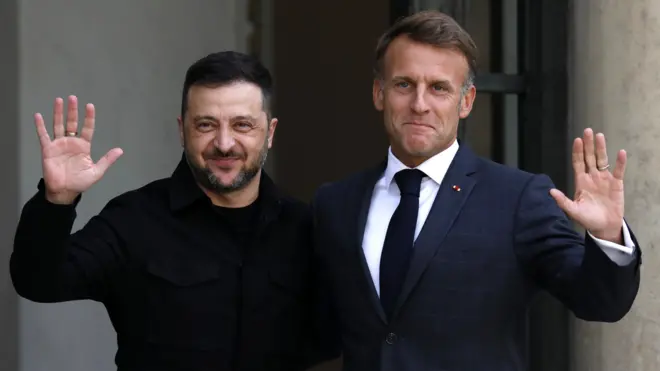 Two men, one in a dark blue suit on the right and another in a black shirt and a beard smile at the cameras while a soldier stands guard on the right