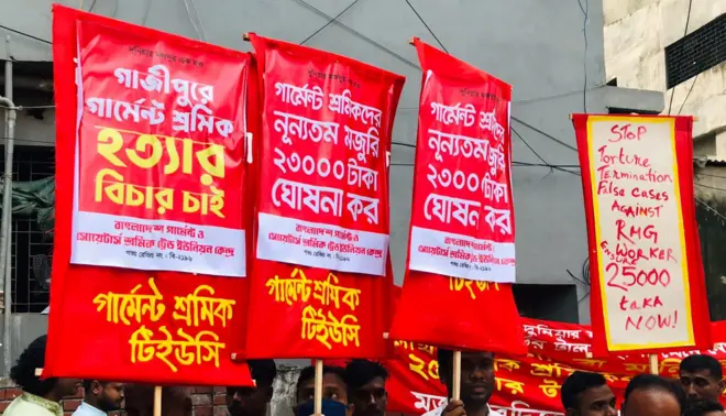 মঙ্গলবার সকালে এরকম প্লাকার্ড নিয়ে নিম্নতম মজুরি বোর্ডের কার্যালয়ের সামনে দাঁড়ান পোশাক শ্রমিকরা