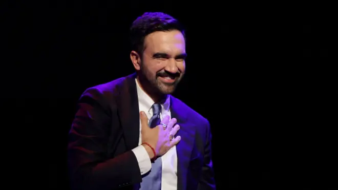 Mamdani celebra su triunfo, sonriente y con una mano en el pecho. Está vestido de terno, con una corbata celeste y el fondo negro. 