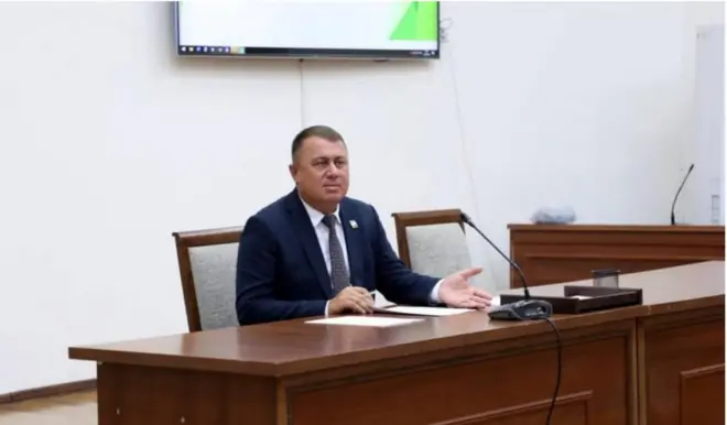 Собиқ Қумқўрғон ҳокими Анвар Норамамтов мажлисда  стол қаршисида ўтириб, микрафонга гапираяпти. У расмда тўқ кўк ранг костюм шимда, оқ куйлак, кўк гакстук таққан.