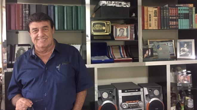 Jorge Perlingeiro sorrindo em frente a estante em casa