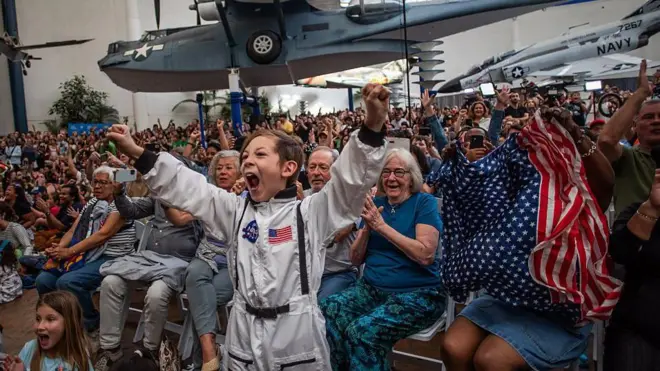 Un niño con disfraz de astronauta celebra entusiasmado el amerizaje de la tripulación de Artemis II, mientras una mujer ondea la bandera de EE.UU. durante una transmisión del evento