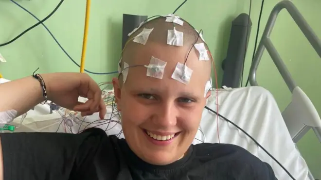 Phoebe Tesoriere sorri sentada em uma cama de hospital passando por um teste de diagnosticado, com diversos fios médicos fixados à sua cabeça raspada