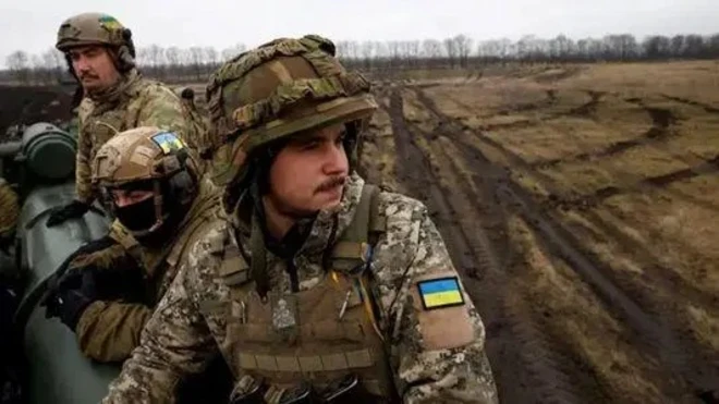 Украинские военные, три человека. Пейзаж ноябрьский либо мартовский, вдалеке виднеются деревья без листьев, на земле следы от техники.