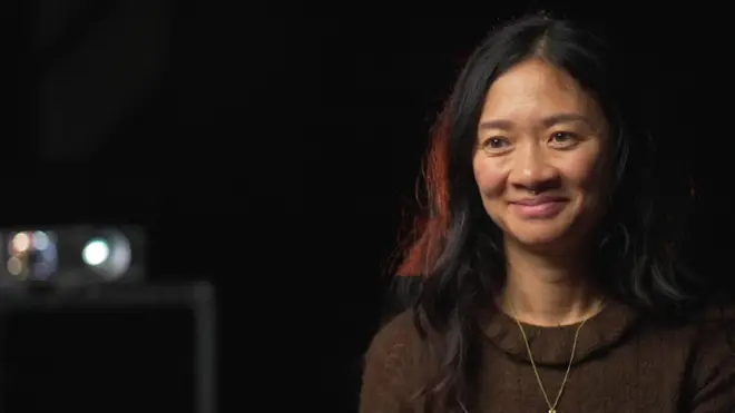 کلویی ژائو با لباس قهوه‌ای و موهای بلند مشکی، لبخند می‌زند