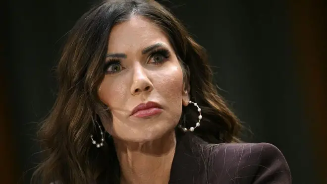 Крупный план: Кристи Ноэм, женщина с длинными каштановыми волосами, с кольцеобразными серьгами в ушах, в коричневом пиджаке, внимательно и настороженно смотрит на кого-то