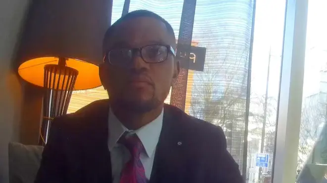 Undercover footage of a man in a suit, wearing glasses, in a hotel lobby.