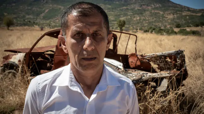 A middle-aged man in white shirt is stood in front of a burned-out car in a field of dried out wild grass, with mountains in the distance