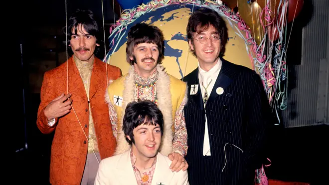 Четверо молодих учасників The Beatles стоять у кольорових сценічних костюмах на тлі великого глобуса. 