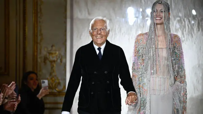 Giorgio Armani podyumda bir mankenle yürüyor.