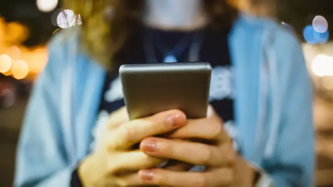 Una mujer sostiene un teléfono móvil. Lleva una camisa celeste. 