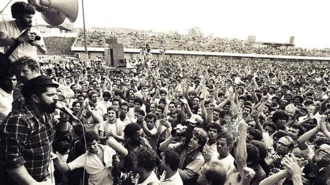 Foto em preto e branco, do início de 1980, com Lula discursando em cima de um pequeno palco para uma multidão em um estádio de futebol.