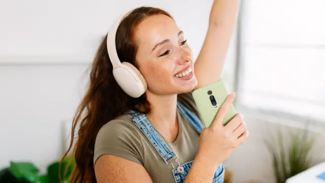 Młoda, uśmiechnięta kobieta trzymająca smartfon jak mikrofon, śpiewająca i ciesząca się chwilą muzyką w domowym zaciszu.