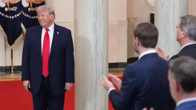 Trump kadrajın soluna doğru bakıyor, yüzünde alaycı bir gülümseme var, önde birkaç kişi onu ayakta alkışlıyor. 