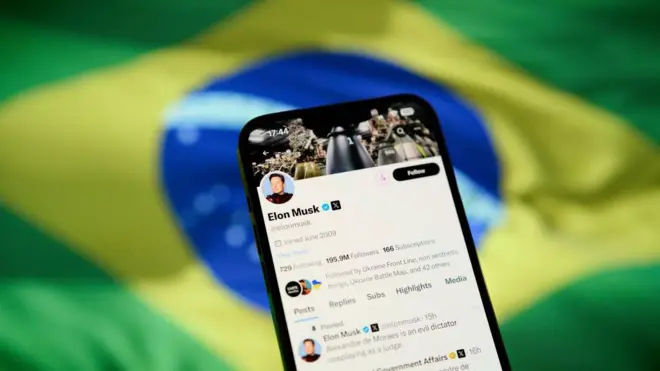 Celular mostrando perfil1xbet é boaElon Musk com bandeira do Brasil ao fundo