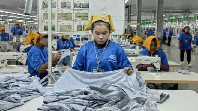 A women wearing a dark blue shirt and a yellow headcloth in a garment factory in Cambodia. She is tending to fabric on a table. Behind her are rows of tables with other similarly dressed women.