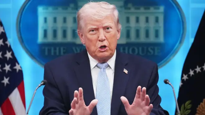 ダークスーツ姿のトランプ氏がホワイトハウスの記者会見場でマイクを前に立ち、両手を前に突き出して口を開いている