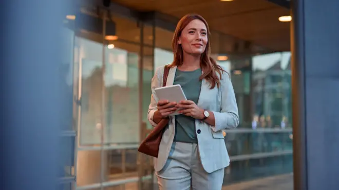Açık renk ceket ve pantalon giyinmiş bir iş kadını elinde bir defter ve omzunda çantasıyla emin bir şekilde yürüyor