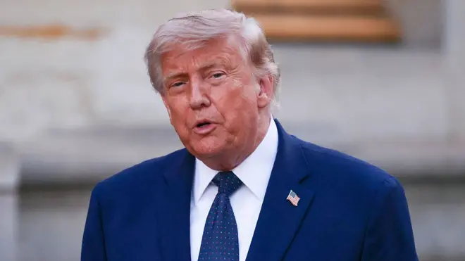 Trump is seen speaking. He is wearing a blue suit and a tie with the American flag as a pin on it 