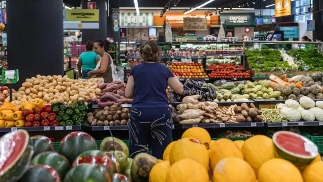 Uma mulhercassino jogos grátiscostas escolhendo legumes na seçãocassino jogos grátisvegetaiscassino jogos grátisum supermercado com outras pessoas passando ao fundo